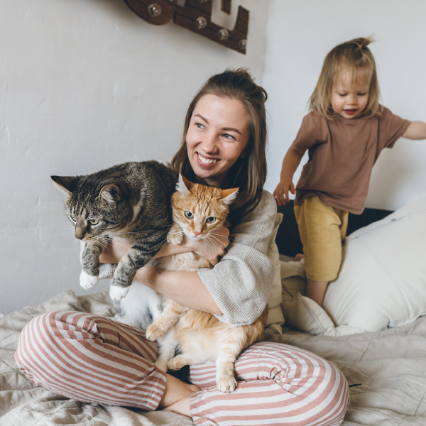 Young woman and her child are sitting on a bed hugging their two cats. One cat is orange, one cat is brown. Pet cremation services.