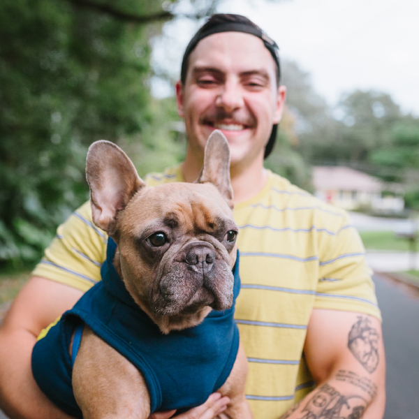 Man wearing backwards cap is smiling and holding his french bulldog. The french bulldog is in a navy blue vest and is close tot he camera. Pet cremation services.